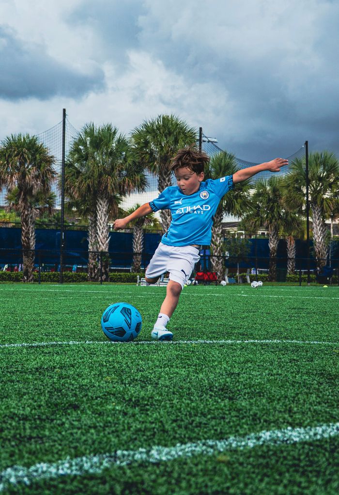 A young boy in a blue jersey actively kicks a soccer ball on a green field amidst palm trees.